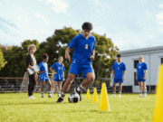 Can High School Sports Win Back the Spotlight? High school soccer players practice on a grassy field at sunset, wearing school uniforms and running drills. The image highlights the spirit of teamwork and school pride amid growing competition from private club sports programs.